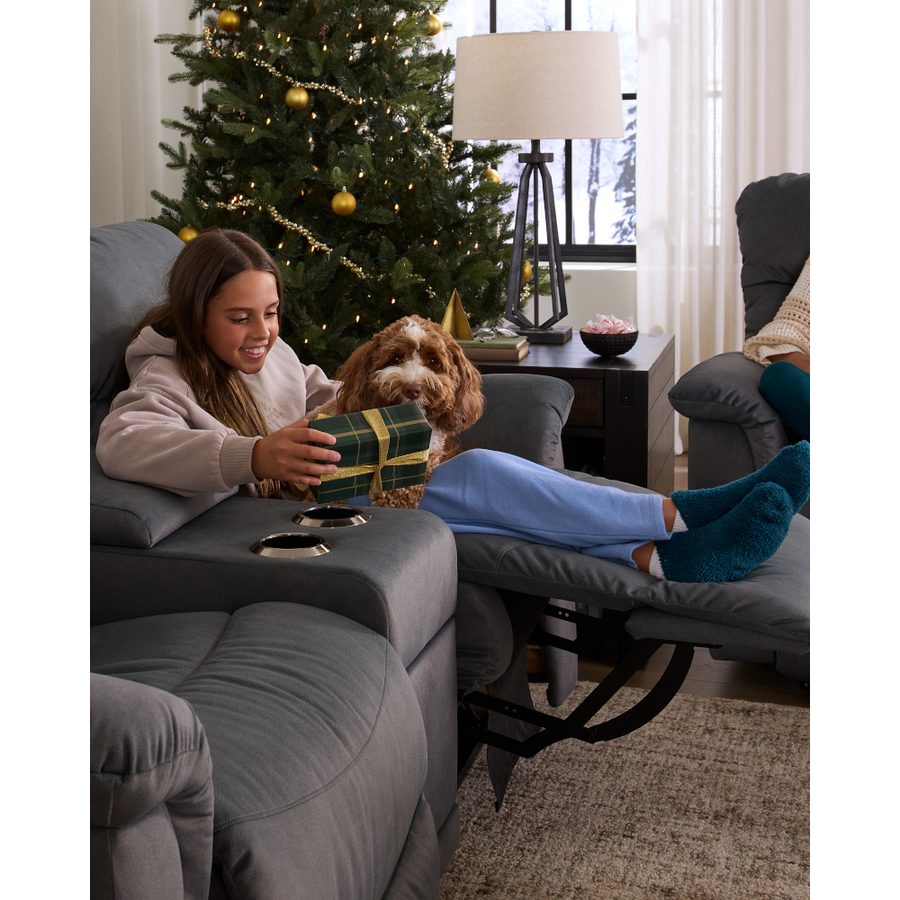 Girl opening a gift with her dog in a gray reclining loveseat beside a Christmas tree
